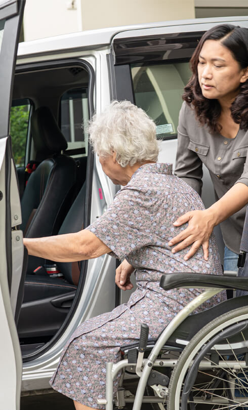 Supreme Cleaning & Care Services photo of a home care worker helping an elderly lady from her wheel chair into the passenger seat of a car.